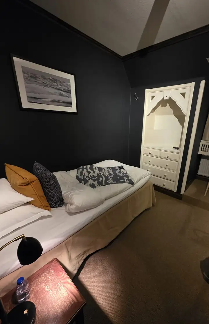 Hotelzimmer mit dunkelblauen Wänden, Bett mit gelbem und gemustertem Kissen, weißer Nische mit Kommode und einem Schwarzweißfoto einer Berglandschaft