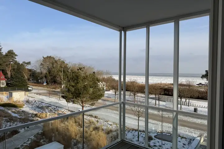 Eckbalkon mit Glasbrüstung im the breeze, davor die verschneite Promenade von Ahlbeck, Dünen und die vereiste Ostsee unter blauem Himmel