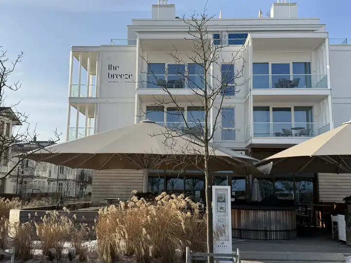 Weiße Fassade des the breeze mit Balkonen aus Glas und dem Schriftzug the breeze, davor Sonnenschirme und Gräser im Schnee