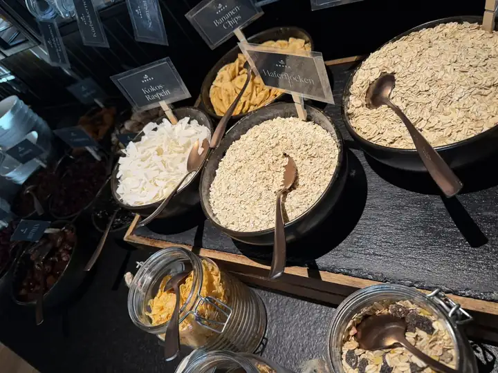 Frühstücksbuffet mit Schalen voller Flocken, Kokosraspeln und Müsli, beschriftet mit kleinen Schildern