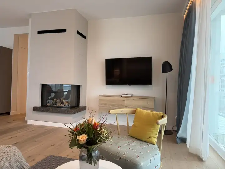 Kamin mit Glas über Eck im Wohnraum des Apartments, davor ein Sessel mit gelbem Kissen und ein Blumenstrauß