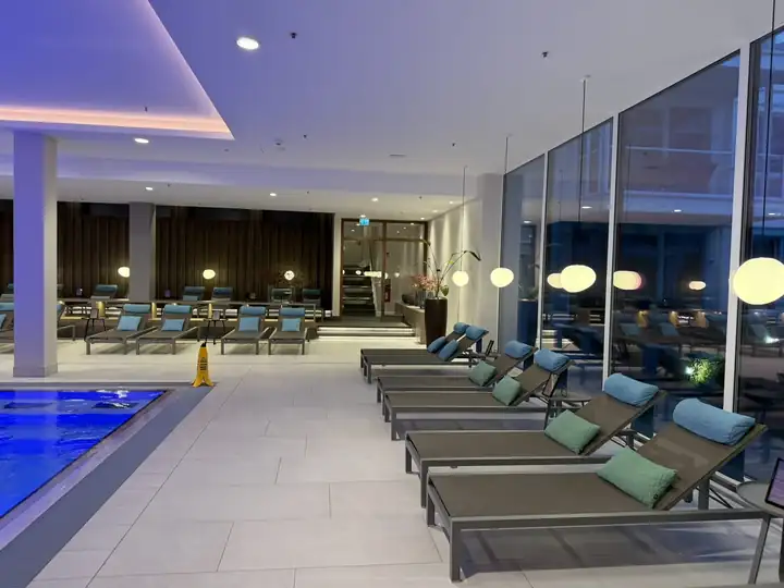 Reihe von Liegen mit blauen und grünen Kissen an der Glasfront des Spa, links das blau beleuchtete Becken