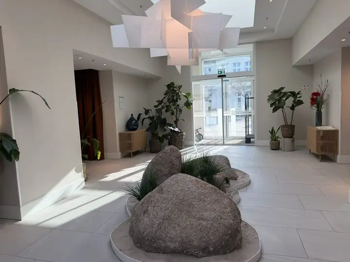 Lobby des the breeze mit Findlingen in einem Beet aus Gräsern, großer weißer Lamellenleuchte, Pflanzen und Glastür