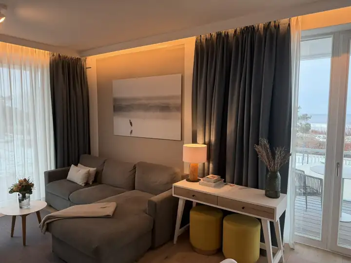 Wohnraum im Apartment mit grauem Sofa, gelben Hockern, weißem Konsolentisch und bodentiefen Fenstern zum Balkon mit Meerblick