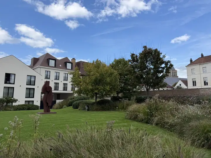 Garten des Dukes' Palace in Brügge mit Rasen, Gräsern, einer Skulptur aus rostrotem Stahl und weißen Hotelflügeln, dahinter ein Kirchturm