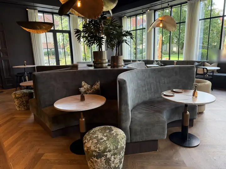 Lounge im Dukes' Palace mit runden Sitznischen aus grauem Samt, Marmortischen, Blumenhockern und Messingleuchten vor Fenstern zum Garten