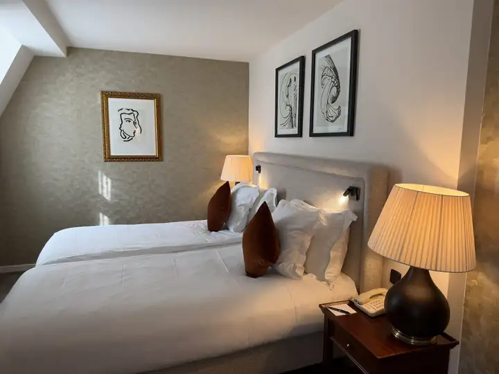 Hotelzimmer unter dem Dach mit zwei Betten, gepolstertem Kopfteil, Tischlampen und Zeichnungen an der Wand