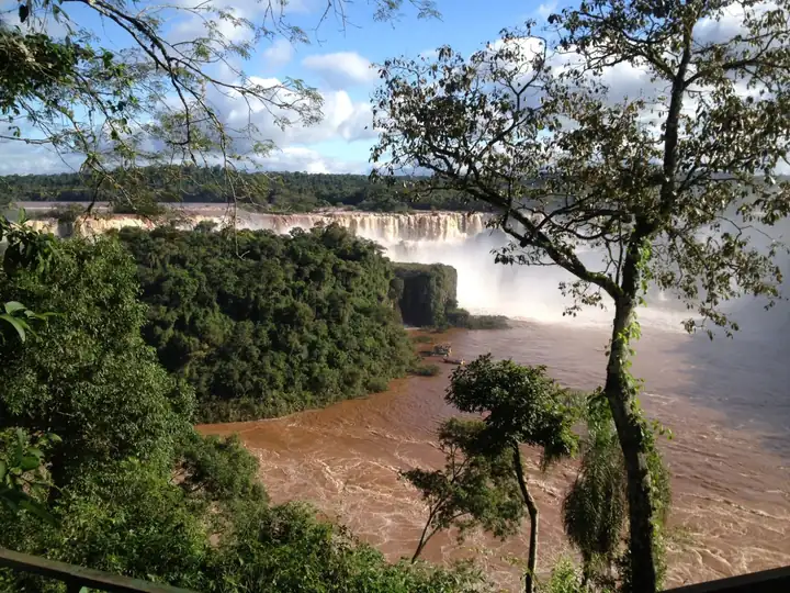 Blick durch Bäume auf die Iguaçu Fälle und den braunen Fluss unter blauem Himmel mit Wolken