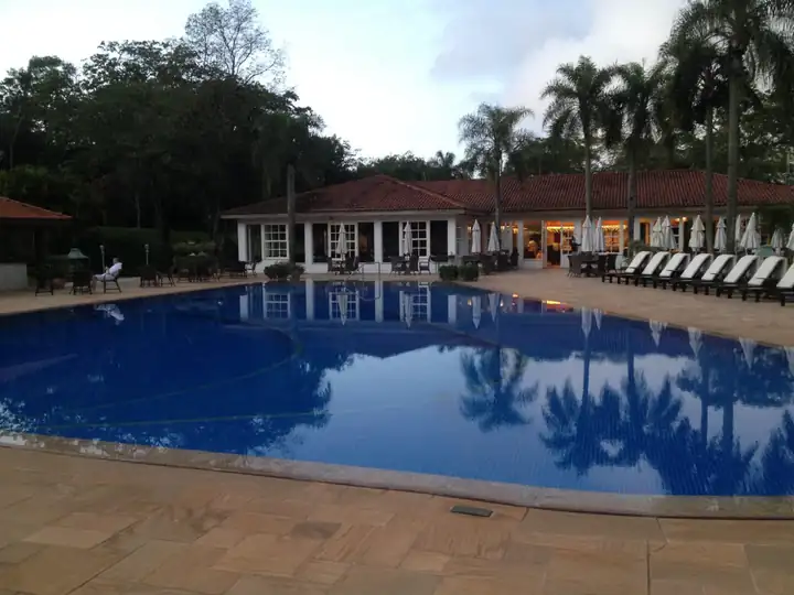 Gebogener Pool mit Spiegelung von Palmen am Abend, dahinter ein Restaurant mit Ziegeldach