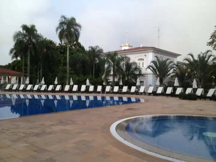 Hotelpool mit Liegen und Palmen vor dem rosa Hauptgebäude unter grauem Himmel