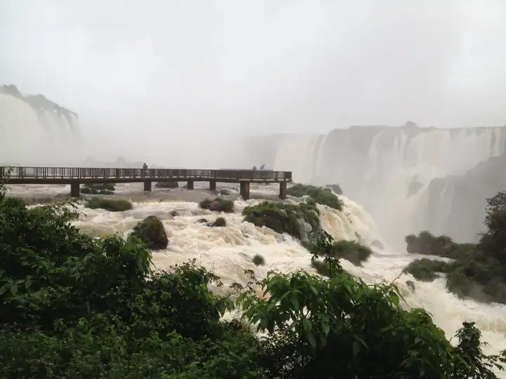 Steg über dem braunen Hochwasser des Iguaçu, dahinter Wasserfälle im Nebel