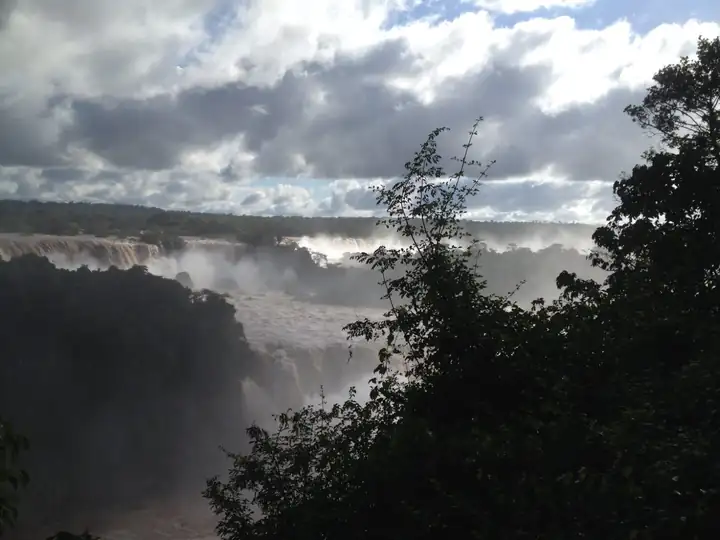 Iguaçu Fälle unter Wolken, im Vordergrund dunkle Bäume, Gischt über dem Fluss