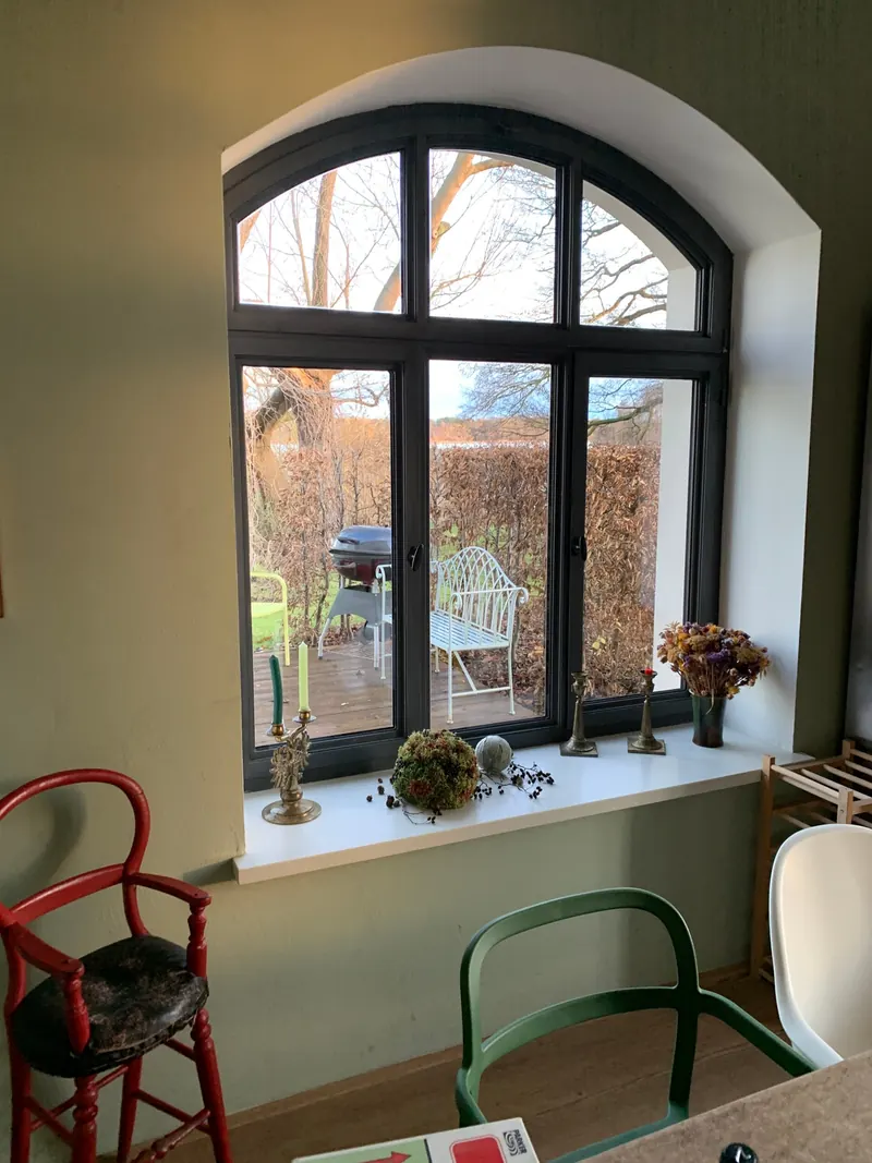 Großes Bogenfenster mit Blick in den Garten, auf dem Fensterbrett Trockenblumen und Kerzenhalter, davor ein roter Hochstuhl und ein grüner Stuhl