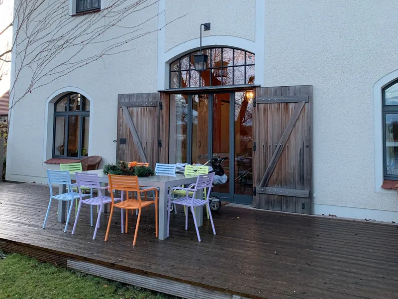Das Haus am See auf dem Eibenhof: weißes Gebäude mit Bogenfenstern und offenen Holzläden, davor eine Terrasse aus Holz mit bunten Stühlen um einen langen Tisch