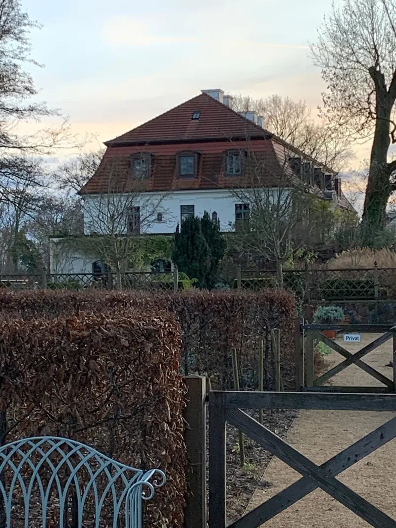 Herrenhaus mit rotem Mansarddach und Gauben hinter einer hohen braunen Hecke, davor ein Holztor und eine verzierte Gartenbank