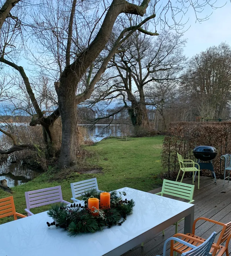 Langer weißer Tisch auf einer Holzterrasse mit Adventskranz und zwei brennenden orangen Kerzen, bunte Stühle, dahinter alte Bäume, Garten und der Scharmützelsee