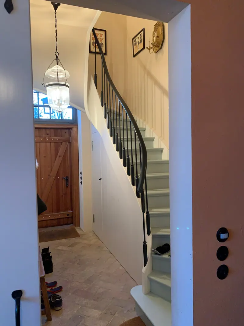 Geschwungene weiße Treppe mit schwarzem Geländer im Flur, unten eine Haustür aus Holz mit Oberlicht, an der Decke eine Laterne