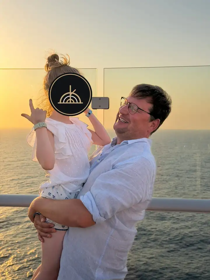 Vater hält seine Tochter an der Reling im Abendlicht, das Gesicht des Kindes ist mit dem GRAND KIN Logo verdeckt