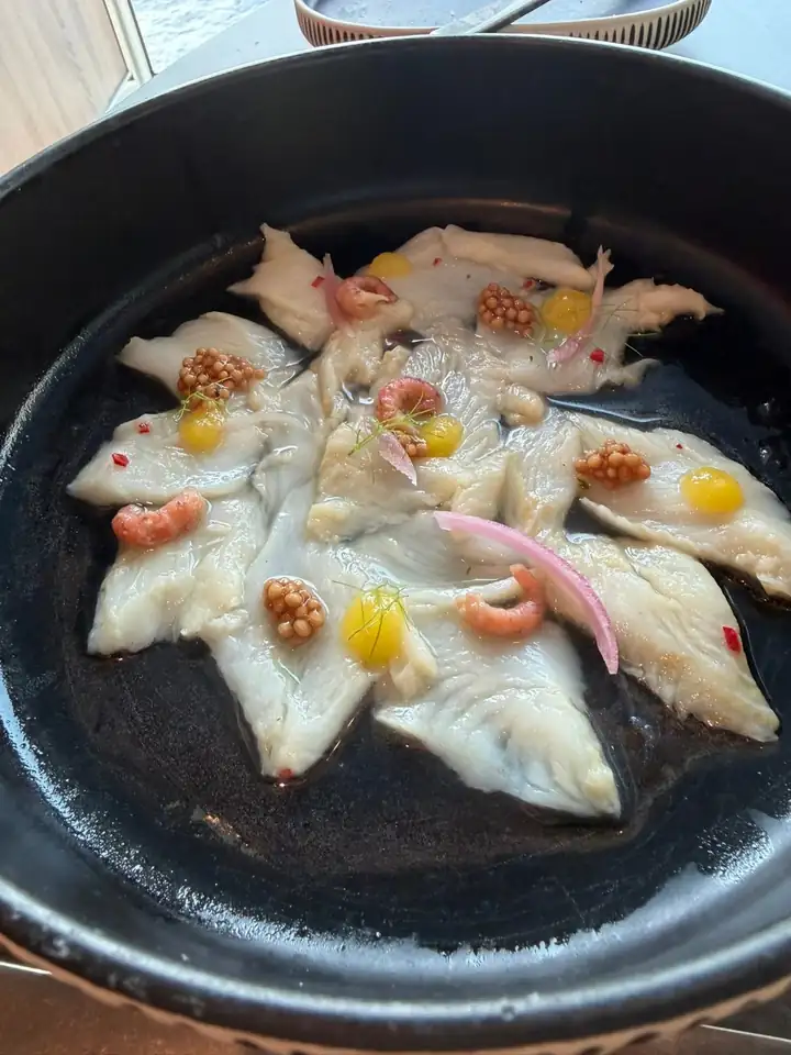 Dunkle Schale mit dünn geschnittenem Fisch, darauf Senfkörner, kleine Garnelen und rote Zwiebel