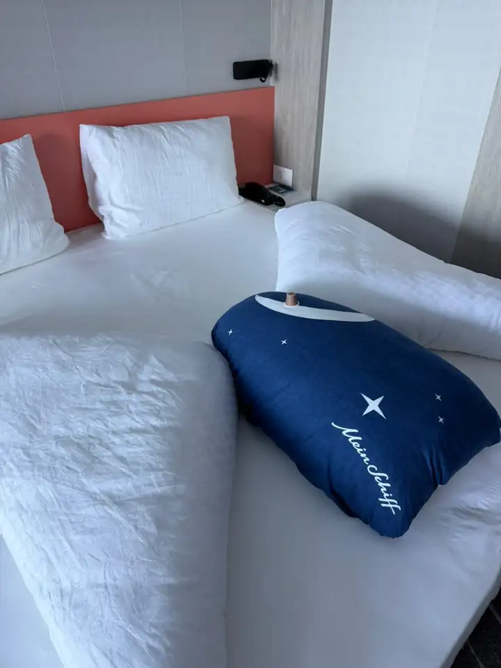 Bett in der Kabine mit weißer Wäsche und einem dunkelblauen Kissen mit Logo