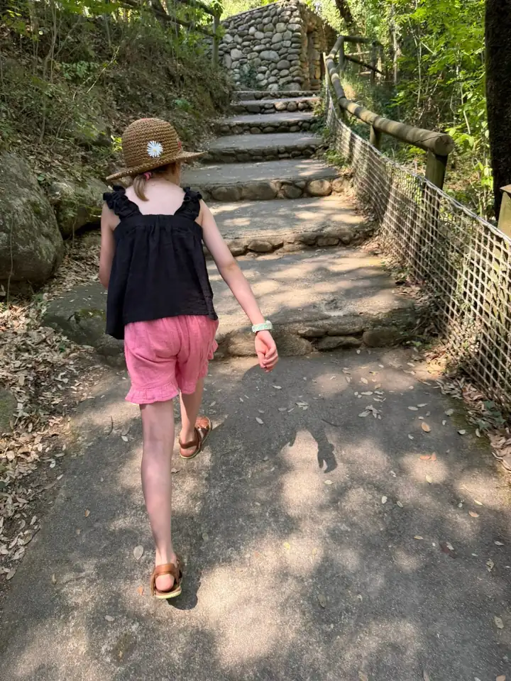 Mädchen mit Strohhut von hinten auf einem Waldweg mit Steinstufen und Holzgeländer