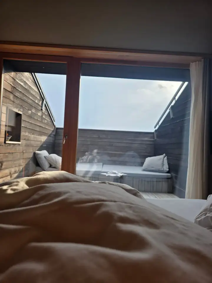 Blick aus dem Bett durch eine große Glastür auf eine Terrasse mit dunklen Holzwänden und einer Liegeinsel mit Kissen