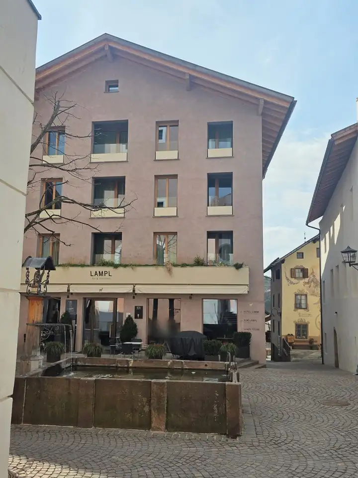 Rosafarbenes Haus mit Holzfenstern am Dorfplatz von Kastelruth, im Erdgeschoss das Lampl Bar und Restaurant mit Terrasse, davor ein steinerner Brunnen