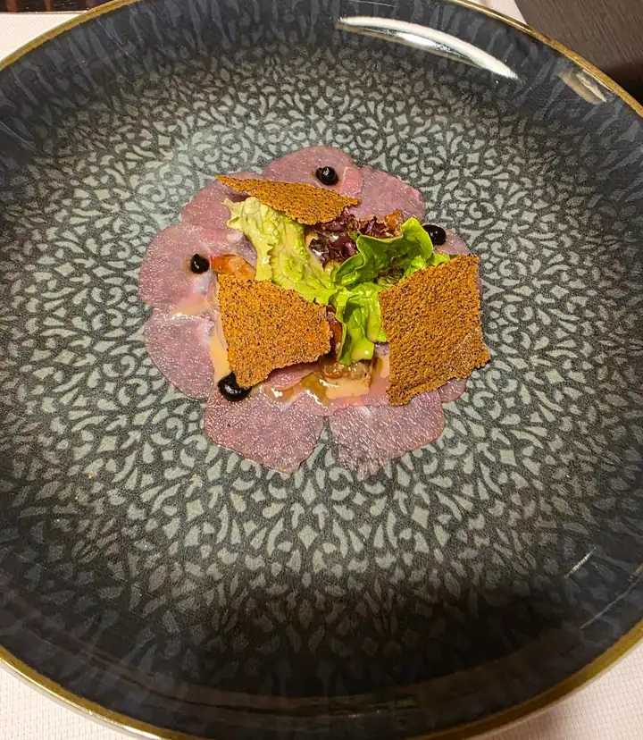 Dünn geschnittenes Fleisch als Carpaccio auf einem dunklen Teller mit Muster, darauf grüner Salat, Knusperblätter und Punkte einer dunklen Sauce