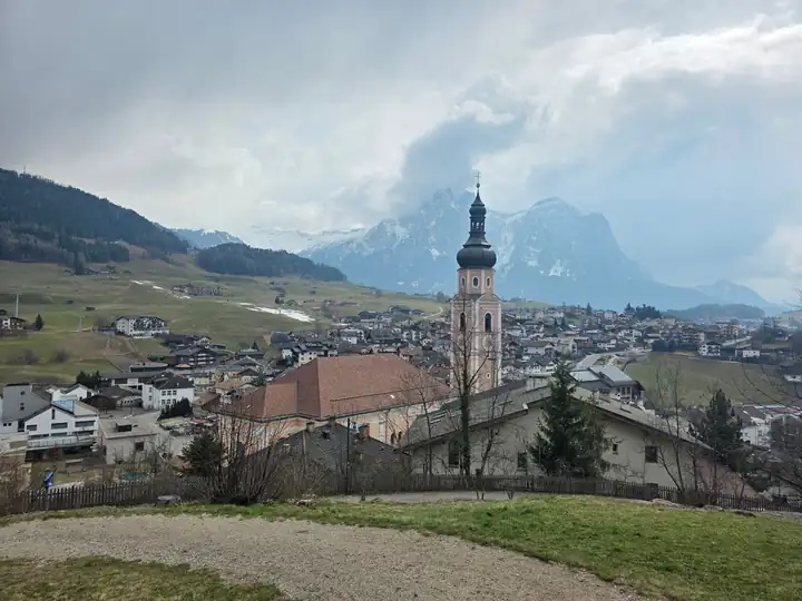 Blick über Kastelruth mit dem Kirchturm, dahinter der Schlern mit Schnee und Wolken und grüne Wiesen am Hang