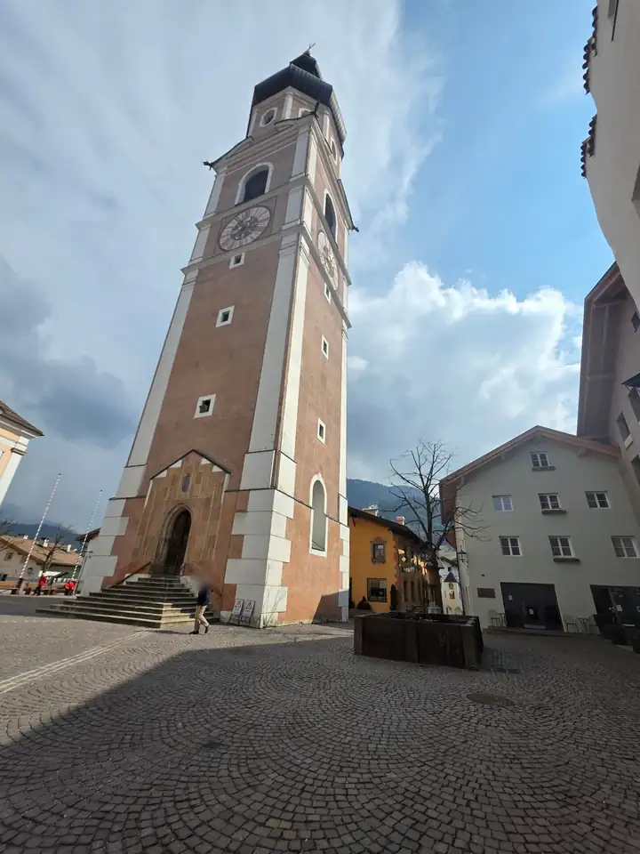 Freistehender Glockenturm der Pfarrkirche von Kastelruth mit Uhr und Zwiebelhaube über einem gepflasterten Platz mit Brunnen