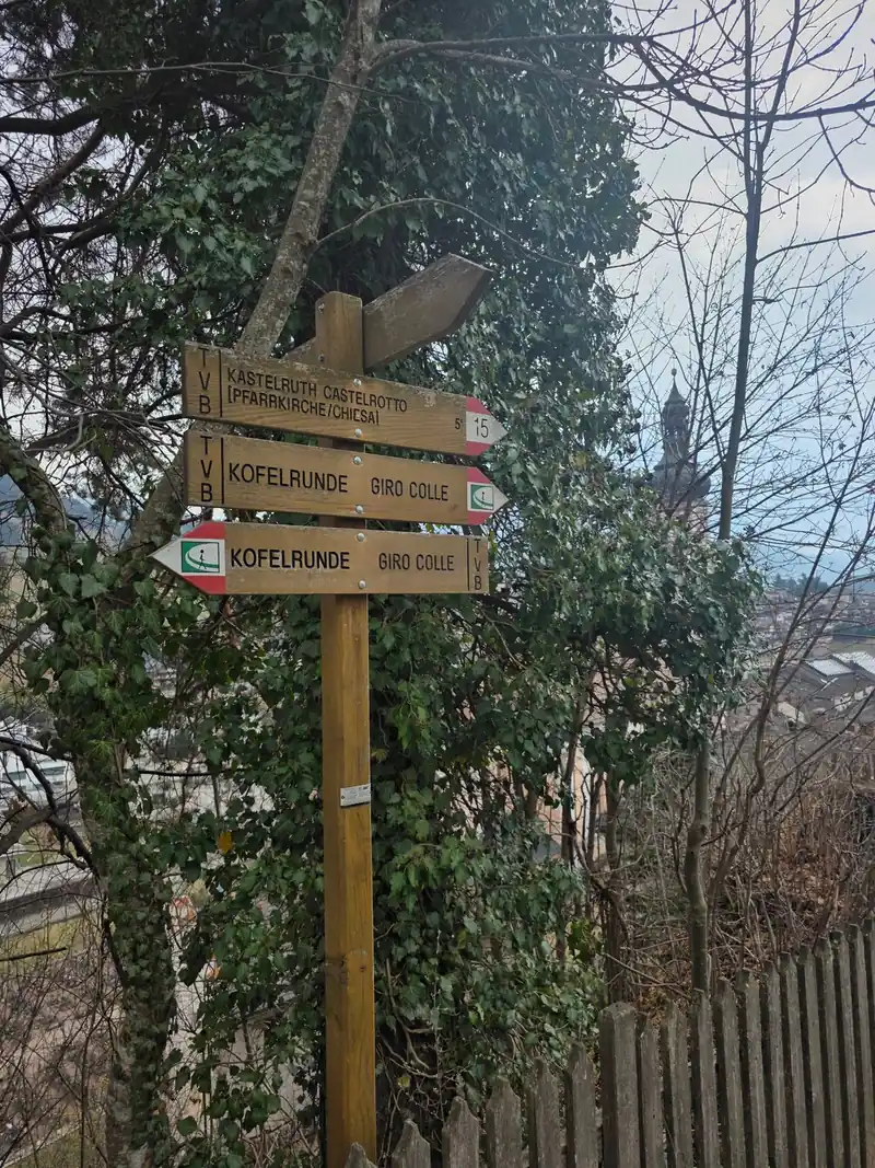 Hölzerner Wegweiser mit den Schildern Kastelruth Pfarrkirche 15 Minuten und Kofelrunde, dahinter Efeu und der Kirchturm