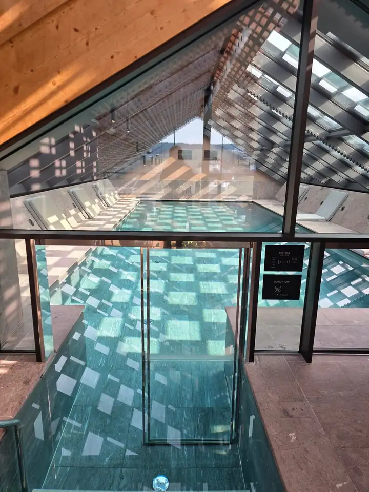 Blick durch eine Glastür auf den Skypool im Dachgeschoss, Holzlamellen werfen ein Lichtmuster auf das türkisfarbene Wasser, an der Seite Liegen