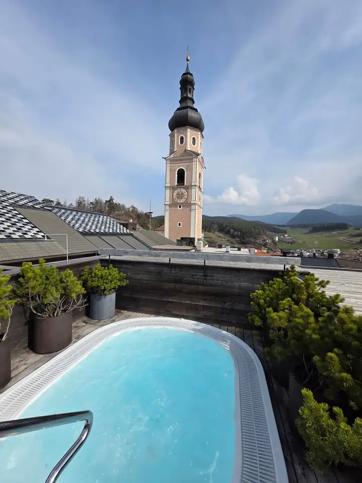 Whirlpool auf der Dachterrasse mit Latschenkiefern in Töpfen, dahinter der Glockenturm von Kastelruth und grüne Hügel