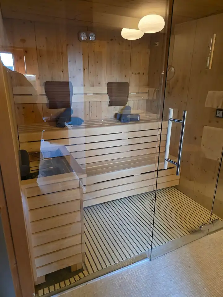 Sauna aus hellem Holz hinter einer Glastür im Zimmer, zwei Liegen mit Kopfstützen und Handtüchern