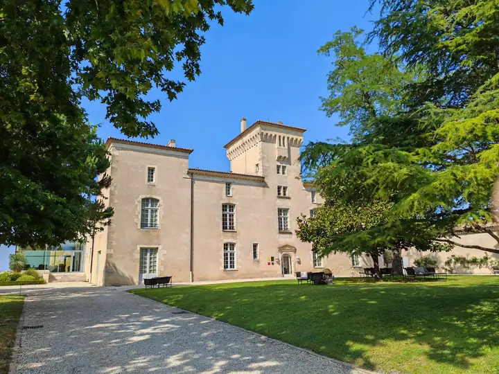 Château Lafaurie Peyraguey mit hellem Steinbau und viereckigem Turm hinter einer Rasenfläche, links und rechts hohe Zedern unter blauem Himmel