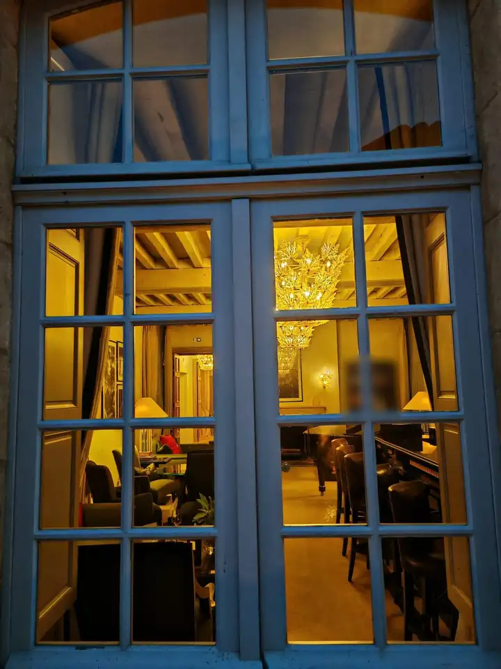 Abends beleuchtetes Sprossenfenster, dahinter ein Salon mit Kronleuchter aus goldenem Kristall und Sesseln