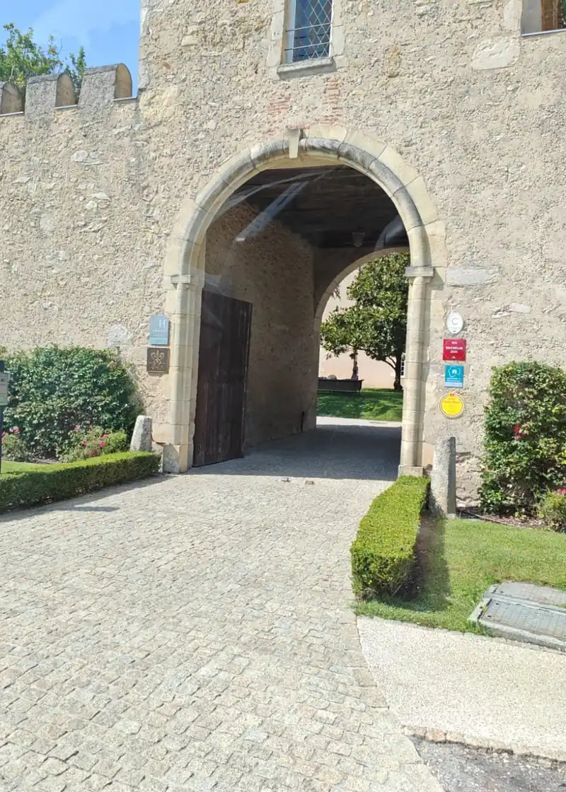 Torhaus aus hellem Naturstein mit Rundbogen und Zinnen, am Pfeiler die Schilder des Hotels, davor ein gepflasterter Weg