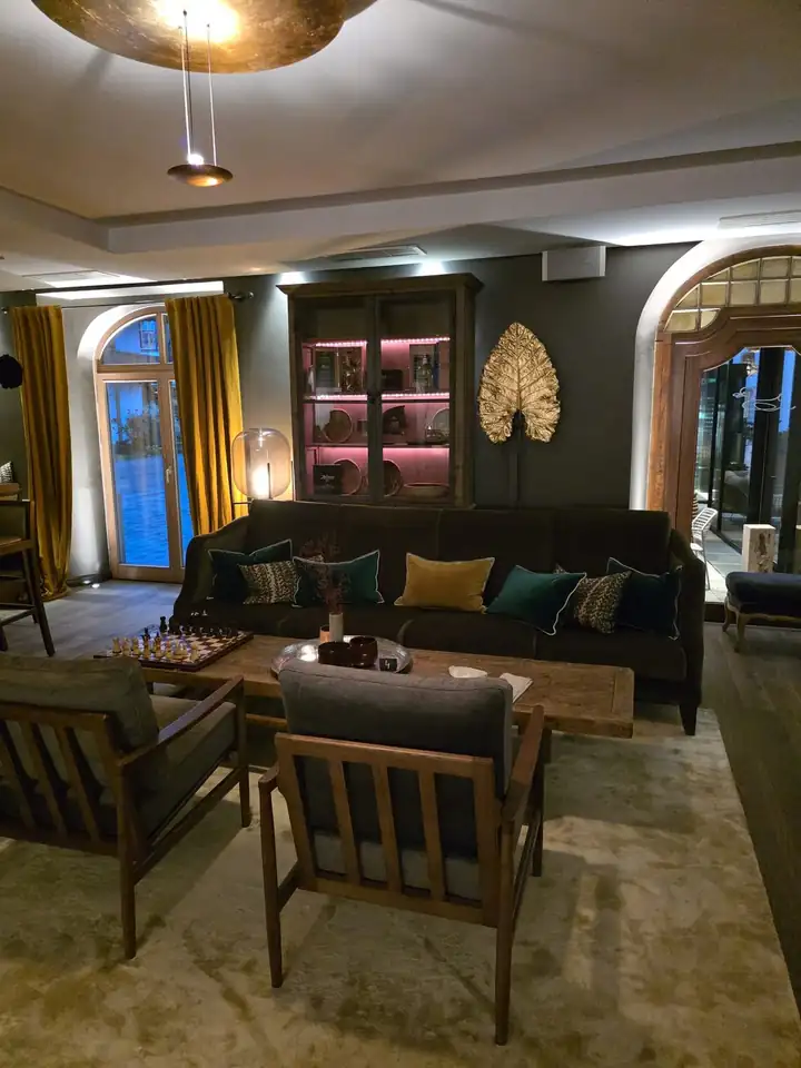 Aufenthaltsraum mit dunklem Samtsofa, Kissen in Grün und Senf, Holzsesseln, einem Schachbrett auf dem Couchtisch und einer Vitrine mit rosa Licht