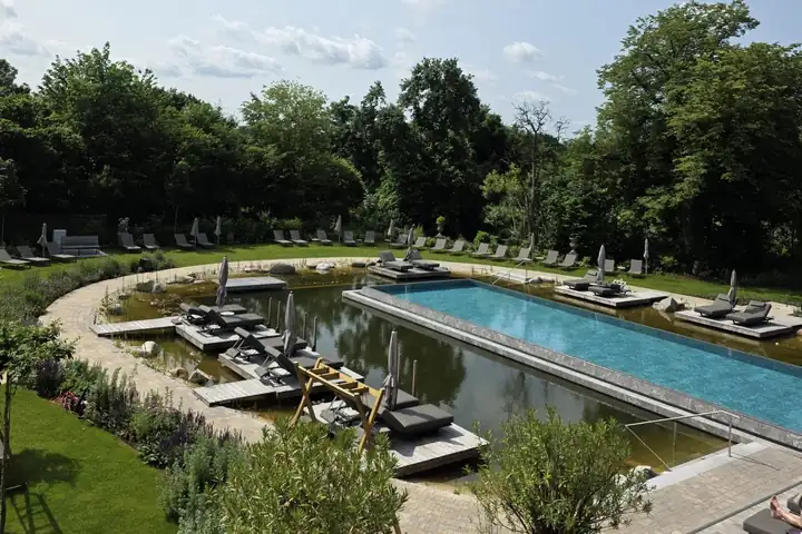 Garten des Mühlbach mit dem langen blauen Thermal Seepool neben einem Badesee mit Holzstegen, Liegen und Schirmen, dahinter hohe Laubbäume
