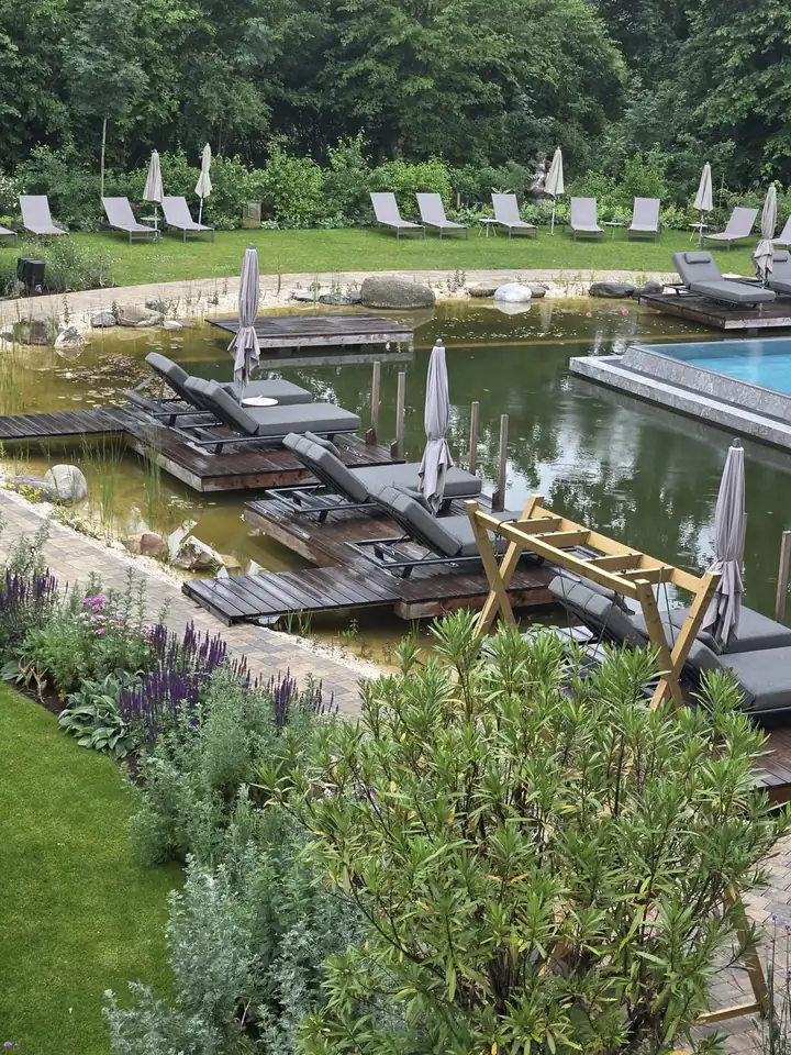 Badesee mit Holzstegen, grauen Liegen und Sonnenschirmen, rechts das Becken des Thermal Seepools, vorne Lavendel und Sträucher