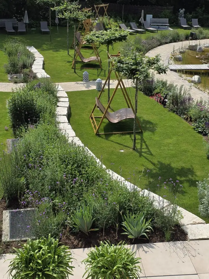 Geschwungener Weg aus Steinplatten durch einen Garten mit Lavendel und Gräsern, auf dem Rasen hängen Schaukelbänke an Holzgestellen