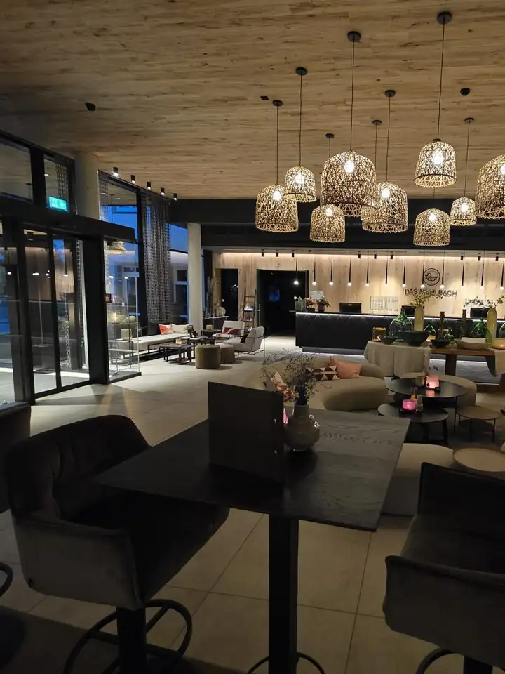 Lobby am Abend mit heller Holzdecke, vielen Hängelampen aus Rattan, Bartischen mit Samthockern und der Rezeption im Hintergrund