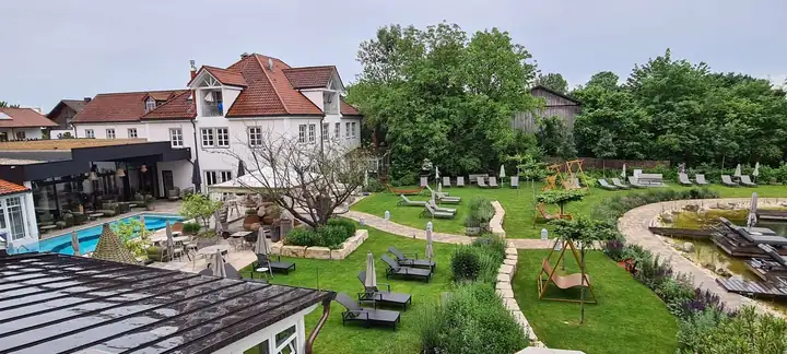 Blick von einem Balkon über den Garten des Mühlbach: links das Stammhaus mit rotem Dach und ein blauer Pool, in der Mitte Rasen mit Liegen und Schaukelbänken, rechts der Badesee mit Holzstegen