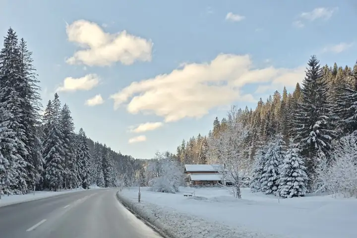Verschneite Landstraße zwischen tief verschneiten Fichten, rechts ein Holzhaus auf einer Wiese, blauer Himmel mit Wolken