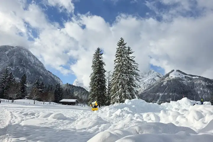 Tief verschneite Wiese bei Achenkirch mit zwei hohen Fichten, dahinter verschneite Berge unter blauem Himmel mit Wolken