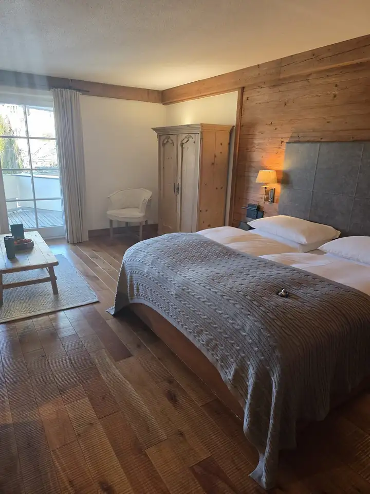 Landhauszimmer mit breitem Holzboden, Doppelbett mit grauer Strickdecke, Bauernschrank aus hellem Holz und Balkontür