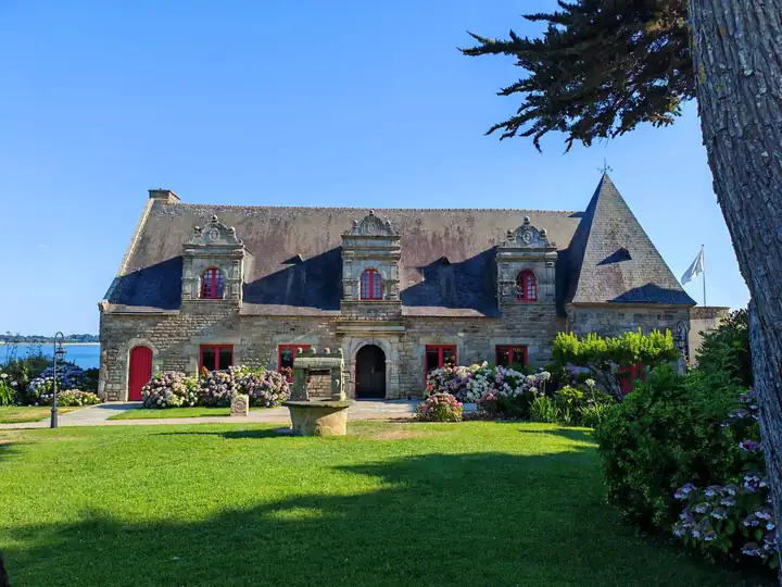 Bretonisches Herrenhaus aus Granit mit verzierten Gauben, roten Türen und Fensterrahmen, davor ein Rasen mit Brunnen und Hortensien