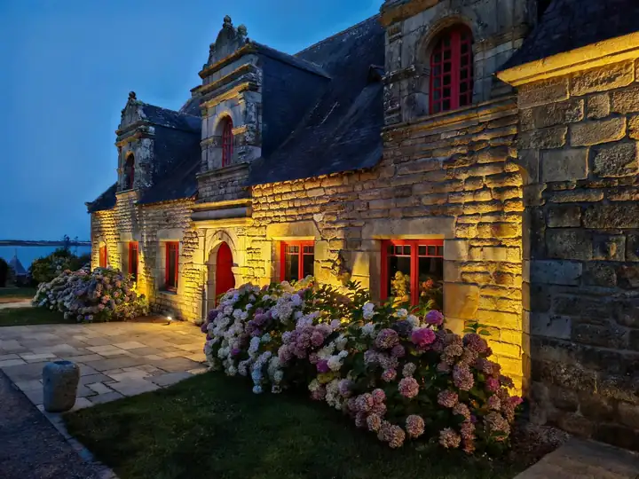 Herrenhaus aus Granit am Abend, von unten angestrahlt, mit roten Fensterrahmen und rosa Hortensien davor