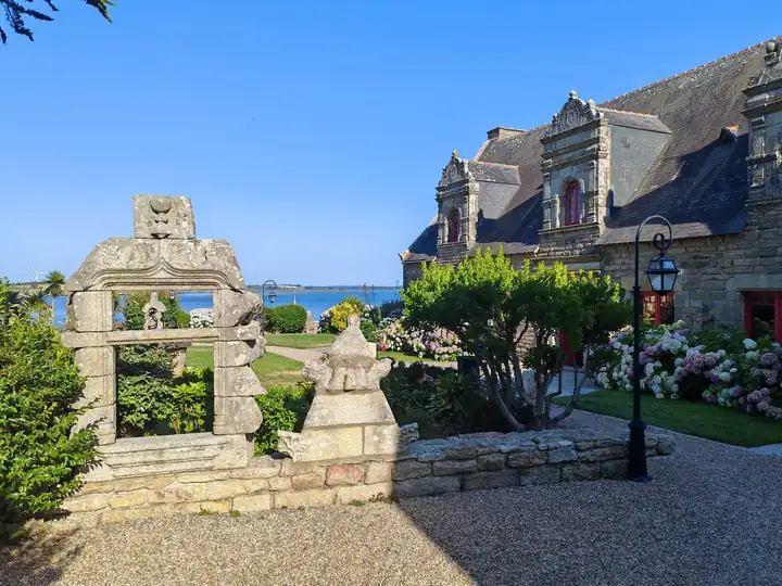Freistehender Fensterrahmen aus verziertem Stein im Garten, rechts ein Herrenhaus, hinten das Meer