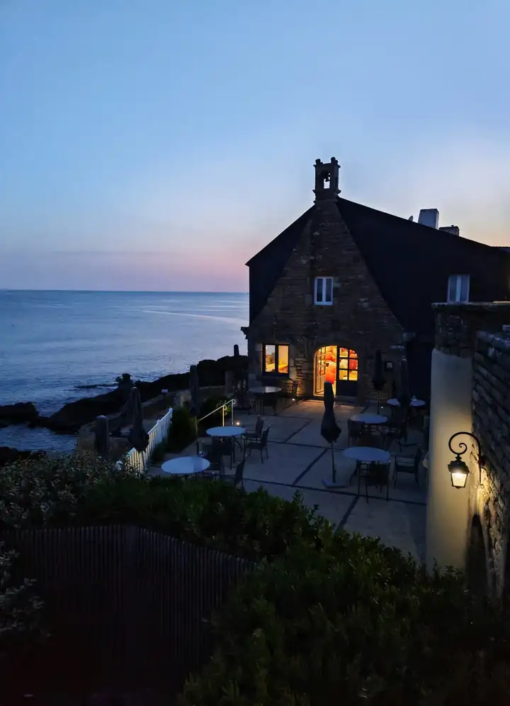 Terrasse mit runden Tischen vor einem Steinhaus mit Glockentürmchen, links der Atlantik und Felsen in der Abenddämmerung